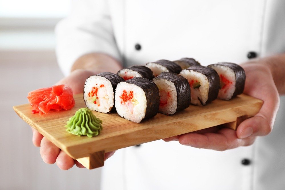 Chef segurando um tabuleiro com oito sushis maki, acompanhados de wasabi e gengibre, em ambiente de cozinha moderna.