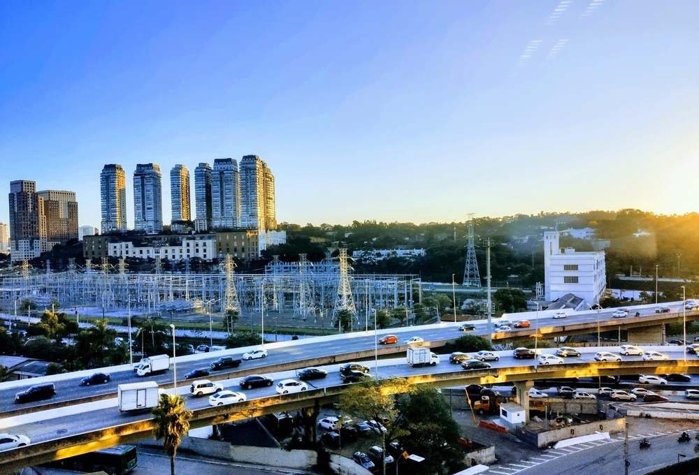 Vista panorâmica de um skyline de edifícios altos modernos ao entardecer, com avenidas movimentadas, árvores e estrutura de transmissão de energia, destacando o crescimento urbano e a vida na cidade.