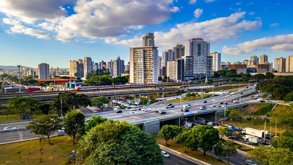 Zona Leste de São Paulo com vários edifícios altos e trânsito movimentado em uma área urbana com árvores no primeiro plano, sob céu claro com nuvens